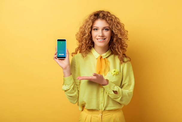 KYIV, UKRAINE - FEBRUARY 4, 2020: happy redhead woman pointing with hand at smartphone with twitter app on screen isolated on yellow  - Photo, Image
