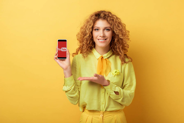 KYIV, UKRAINE - FEBRUARY 4, 2020: happy redhead woman pointing with hand at smartphone with youtube app on screen isolated on yellow  - Foto, Imagem