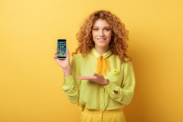 KYIV, UKRAINE - FEBRUARY 4, 2020: happy redhead woman pointing with hand at iphone isolated on yellow  - Foto, Imagem