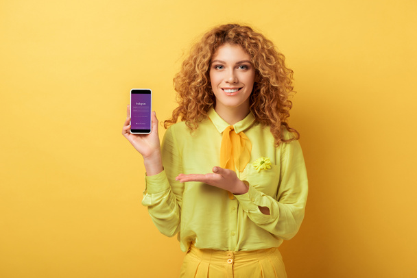KYIV, UKRAINE - FEBRUARY 4, 2020: happy redhead woman pointing with hand at smartphone with instagram app on screen isolated on yellow  - Fotografie, Obrázek
