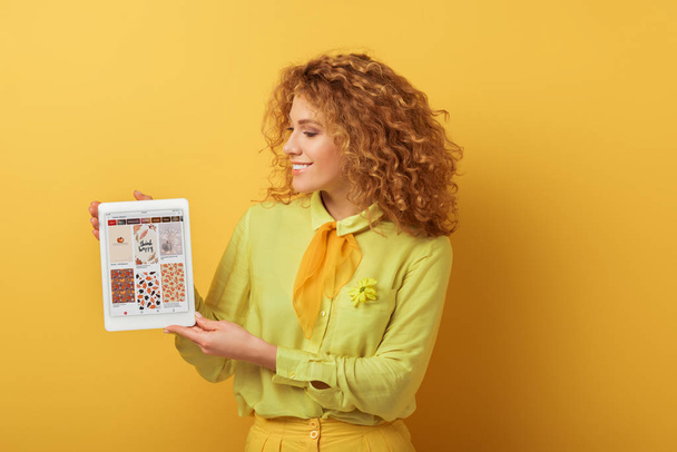 KYIV, UKRAINE - FEBRUARY 4, 2020: cheerful redhead woman holding digital tablet with pinterest app on yellow  - Photo, image