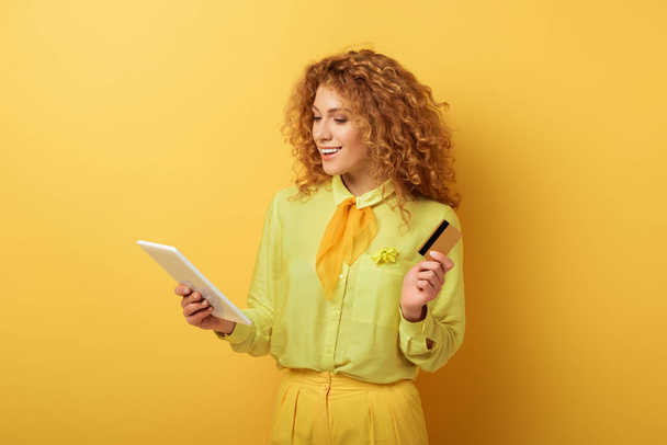 cheerful redhead woman holding digital tablet and credit card on yellow  - Foto, Imagen