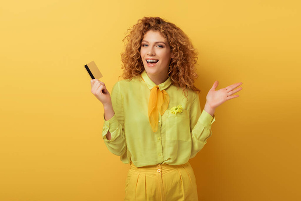 opgewonden roodharige vrouw met credit card en gebaren op geel  - Foto, afbeelding