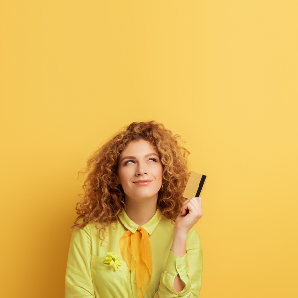 smiling redhead woman holding credit card while thinking on yellow  - Foto, Imagem
