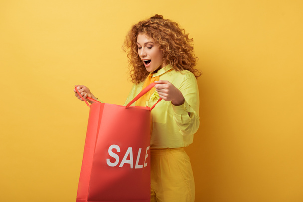 surprised redhead woman looking at shopping bag with sale lettering on yellow  - Photo, Image