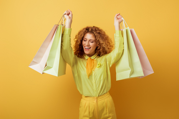 happy redhead woman with closed eyes holding shopping bags on yellow  - Фото, зображення