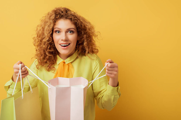 excited redhead girl looking at camera and holding shopping bags on yellow  - Foto, immagini