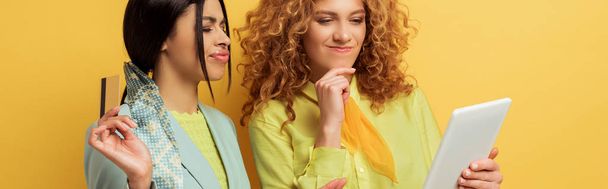 panoramic shot of confused redhead girl using digital tablet near attractive african american woman holding credit card isolated on yellow  - Fotografie, Obrázek