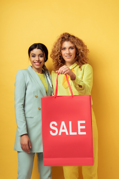 happy redhead girl holding shopping bag near smiling african american woman on yellow  - Photo, Image