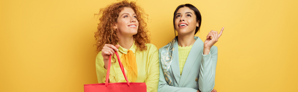 panoramic shot of happy redhead girl holding shopping bag near smiling african american woman pointing with finger isolated on yellow  - Foto, immagini