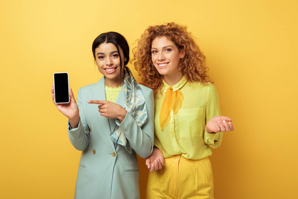 счастливая африканская американка, указывающая пальцем на смартфон с пустым экраном рядом с рыжей девушкой на желтом
  - Фото, изображение