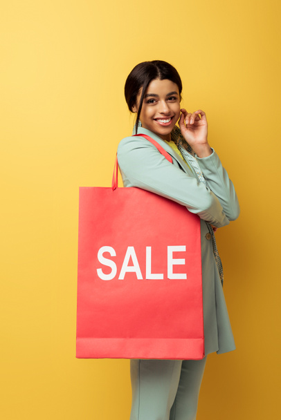 positive african american woman holding shopping bag with sale lettering and smiling on yellow  - Fotografie, Obrázek