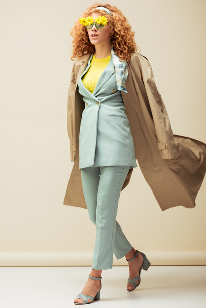 stylish redhead woman in sunglasses with flowers and trench coat walking on beige  - Fotoğraf, Görsel