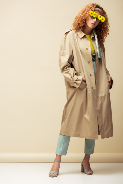 trendy redhead woman in trench coat and sunglasses with flowers standing with hands in pockets on beige  - Photo, Image