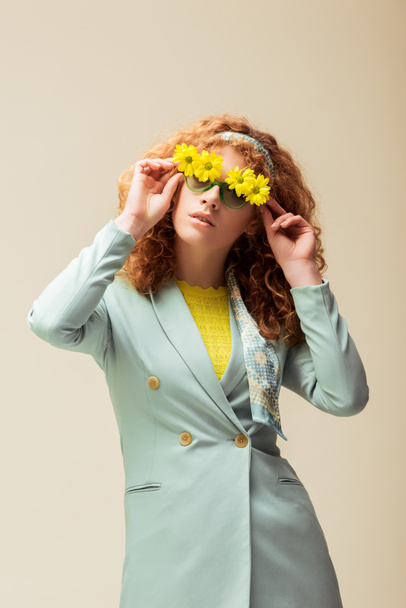 stylish redhead woman in suit touching sunglasses with flowers while posing isolated on beige  - Photo, Image