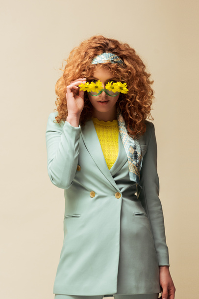 curly and redhead woman in suit touching sunglasses with flowers while posing isolated on beige  - Photo, Image