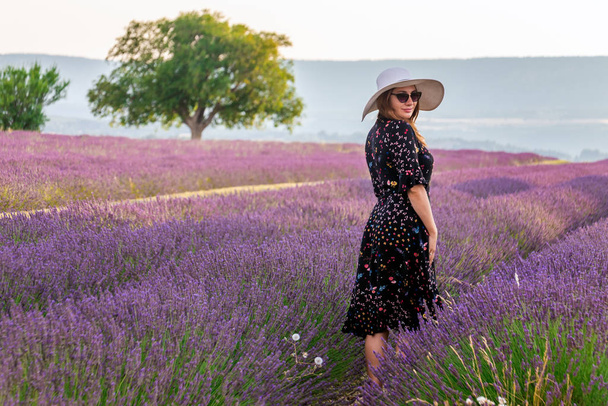 Κορίτσι σε ένα φόρεμα με λουλούδια και μεγάλο λευκό καπέλο στα χωράφια λεβάντας στην Προβηγκία, Valensole, Γαλλία. Γραφική καλοκαιρινή φύση τοπίο και τη γεωργία περιοχή. Δημοφιλής προορισμός ταξιδιού. - Φωτογραφία, εικόνα