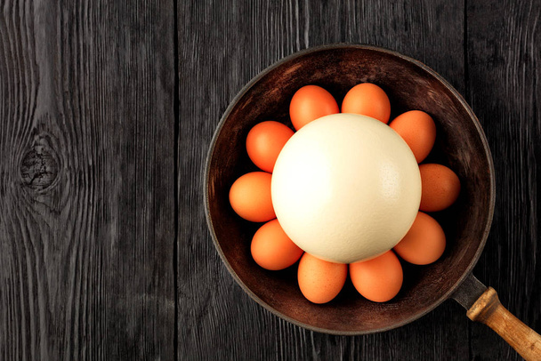 Velké pštrosí vejce obklopeno kuřecími vejci ve staré litinové pánvi, která stojí na starém černém dřevěném povrchu, nízký klíčový obraz, horní pohled, kopírovací prostor. - Fotografie, Obrázek