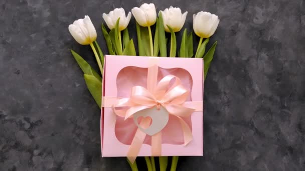Bílé tulipány květiny s růžovou dárkovou krabičkou a štítkem srdce na tmavém pozadí ploché lay.4k video 8 Březen Happy Womens Den matek.Jarní kytice, stuha luk dárek, ženský blog reklama - Záběry, video