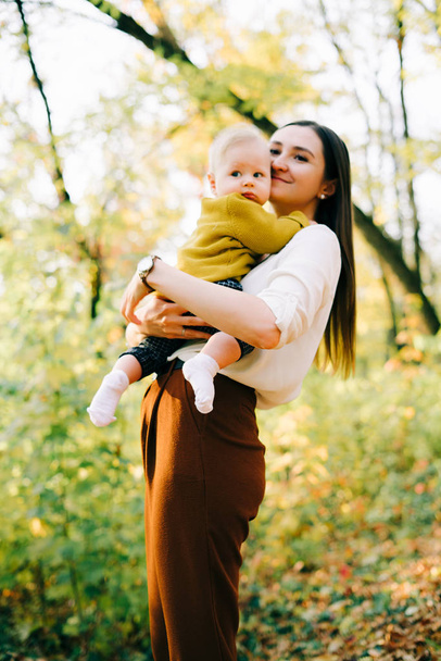 幼い母親は夕日の光の中で秋の木々の間に小さな幸せな息子を抱いています - 写真・画像