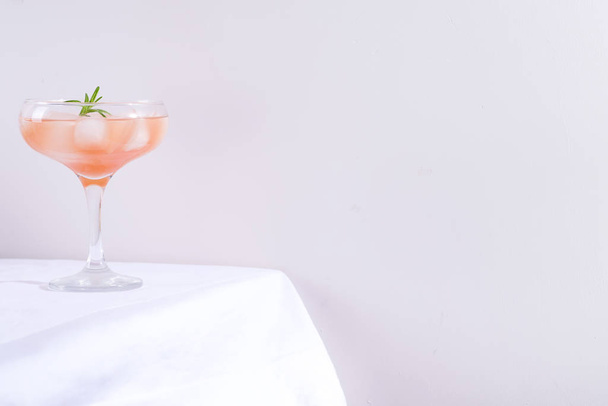 Coquetel rosa com alecrim e gelo em vidro em uma toalha de mesa branca na mesa
 - Foto, Imagem