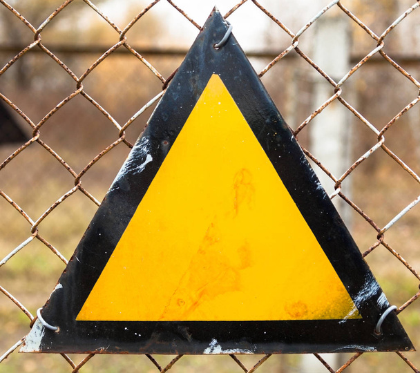 Triangle warning sign on the metal mesh fence. Basic warning sign triangle with a black border and amber background. - Photo, Image