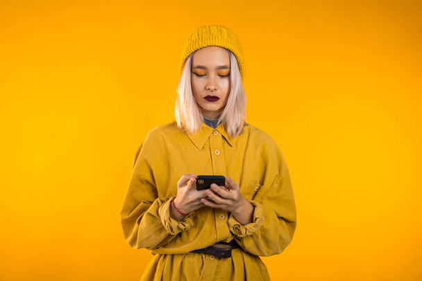 Girl with white hair and unusual make-up using smartphone or surfing internet on yellow studio background. Modern technology - apps, social networks - 写真・画像