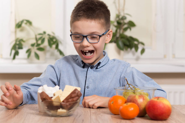 Мальчик в очках сидит за столом и смотрит на сладости со счастливым выражением лица. Рядом с ним лежат мандарин, яблоко и виноград фрукты
. - Фото, изображение
