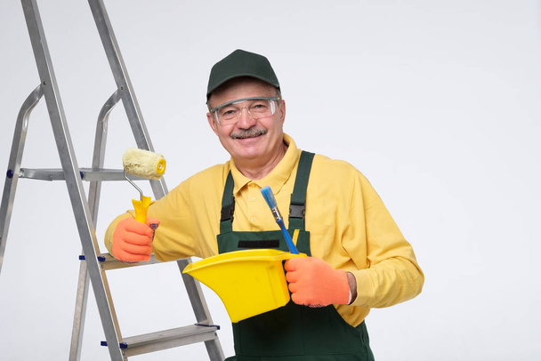 Pittore uomo anziano al lavoro con un rullo di vernice, in piedi vicino alla scala
. - Foto, immagini