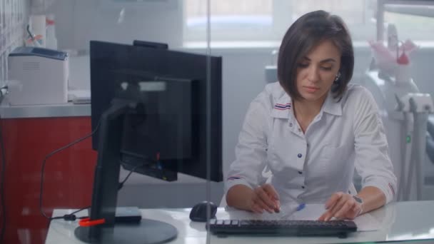Професійна жінка-лікар в лабораторному пальто, друкуючи на комп'ютері з монітором, що сидить за скляною стіною в офісі
. - Кадри, відео