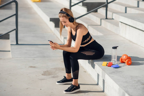 A beautiful fitness girl in grey sportswear uses smartphone and listens to music at the stadium after workout. Sports and healthy concept. - 写真・画像