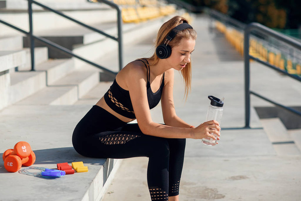 Young caucasian woman drinks water and listen to music after training. - Foto, afbeelding