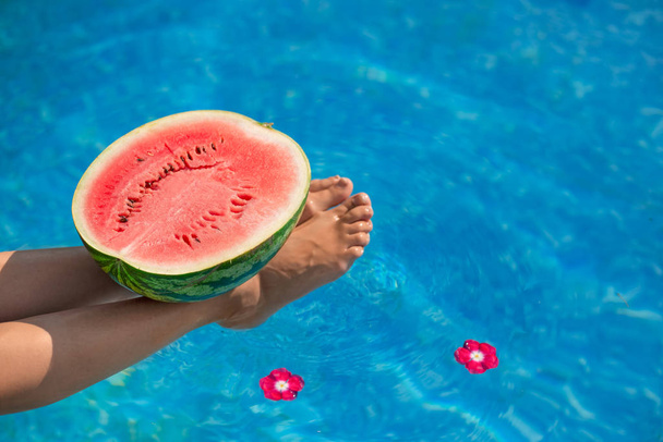 Watermelon in pool on woman slim legs in summer holiday time. Instagram tropical beach luxury lifestyle. Woman relaxing, watermelon and spa flowers in resort blue swimming pool. Foot spa and skin care. - Photo, image