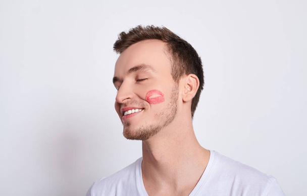 Happy loving handsome young charming handsome white guy with green eyes in a white T-shirt, imprinted with red lipstick, enjoys a kiss, a dream of a beloved, isolated on a white background. - Foto, afbeelding