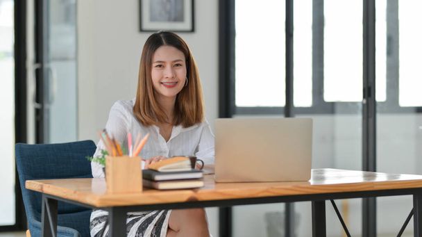 Oříznutý záběr krásné usmívající se podnikatelky pracující při sezení na svém pracovišti v pohodlné kancelářské místnosti - Fotografie, Obrázek