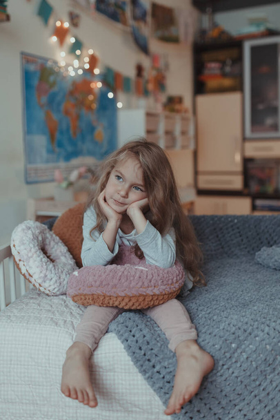 glücklich kleines Mädchen spielt auf dem Sofa im Zimmer, um sie herum sind Plüsch-Donuts und ein kariertes, drinnen - Foto, Bild