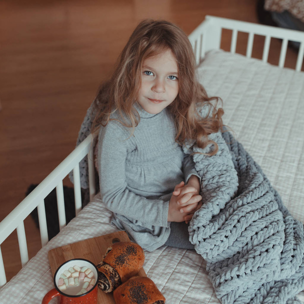 Mooi klein meisje drinkt cacao uit een rode mok op haar bed in een gezellige kamer - Foto, afbeelding