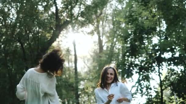 Two attractive women are jumping and smiling at the camera on a warm sunny summer day. Fun photo shoot for couple of cheerful beautiful casual girls in a cozy city park among trees. Video for memory. - Πλάνα, βίντεο