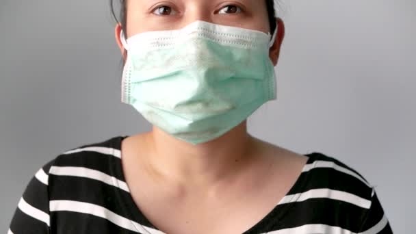 Aziatische jonge vrouw met gezichtsmasker ter bescherming tegen luchtverontreiniging of virusepidemie op grijze achtergrond. Begrippen luchtverontreiniging, stofallergieën en gezondheidszorg. - Video