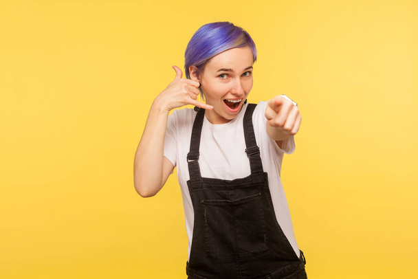 Hey you call me! Portrait of energetic hipster girl with violet short hair in denim overalls showing phone gesture and pointing camera, connection communication concept. yellow background, studio shot - Foto, immagini
