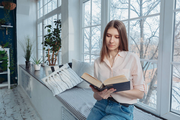 A girl is reading a book by the window. Indoor plants are visible in the background. High quality photo - Fotó, kép