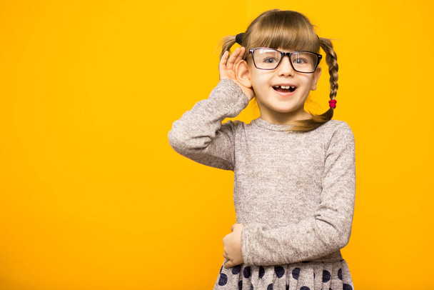 A young child girl journalist in glasses with funny pigtails listening to something holding his hand to ear isolated on yellow background. - Photo, Image