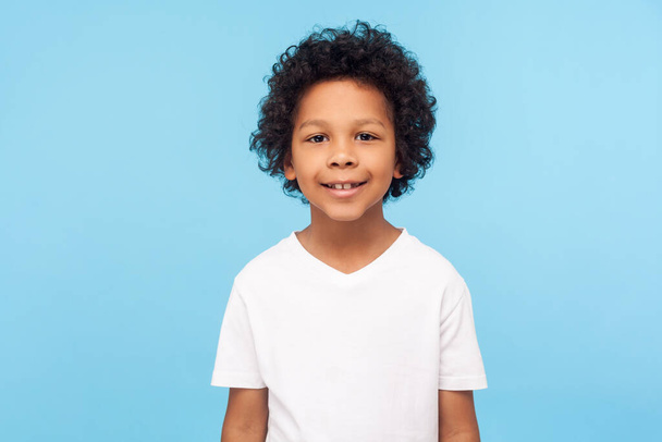 Portrait of cheerful little boy with curly hair in T-shirt smiling funny and carefree, showing two front teeth, healthy happy child, positive emotions. indoor studio shot isolated on blue background - 写真・画像