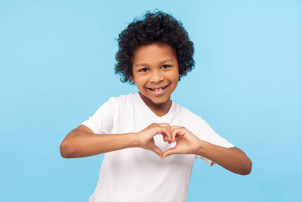 Portrait of smiling cheerful little boy with curly hair in white T-shirt showing heart shape with fingers, expressing innocent childhood love, affection. indoor studio shot isolated on blue background - Photo, Image