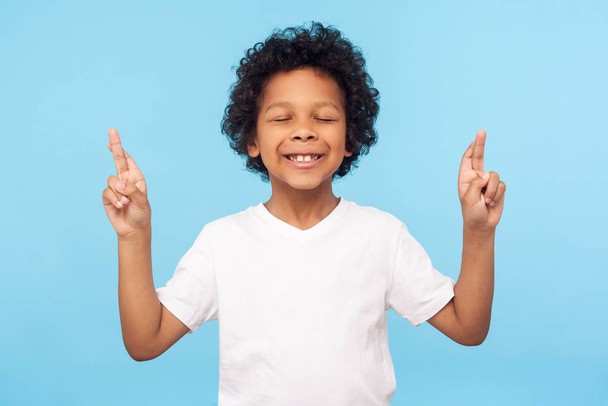Dromen en hoop uit de kindertijd. Portret van een schattig jongetje met gesloten ogen en kruisende vingers, wensend op geluk, wachtend op een wonder. indoor studio shot geïsoleerd op blauwe achtergrond - Foto, afbeelding