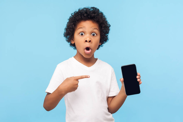 Wow awesome mobile app. Amazed excited cute little boy pointing at cell phone and looking at camera with shocked face, child surprised by telephone, online service. indoor studio shot blue background - Foto, Imagem