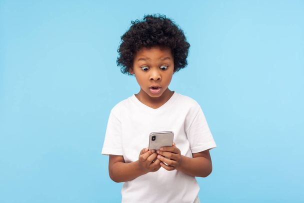 Astonished cute little boy with curly hair reading message on smartphone and expressing amazement shock, surprised by mobile application, using cellphone. studio shot isolated on blue background - Foto, imagen