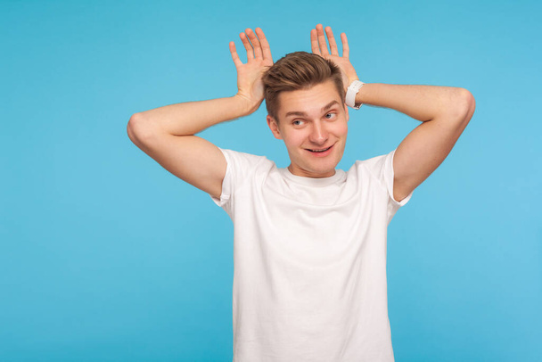 I'm rabbit! Portrait of funny carefree man in casual white t-shirt making bunny gesture with hands on head and smiling with humorous comical expression. indoor studio shot isolated on blue background - Foto, Bild