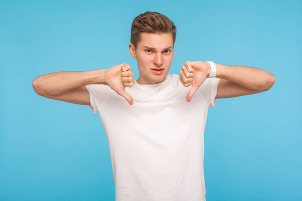 No, don't like this! Portrait of dissatisfied man in white t-shirt looking displeased and showing thumbs down gesture, expressing disagreement, critique. indoor studio shot isolated on blue background - 写真・画像