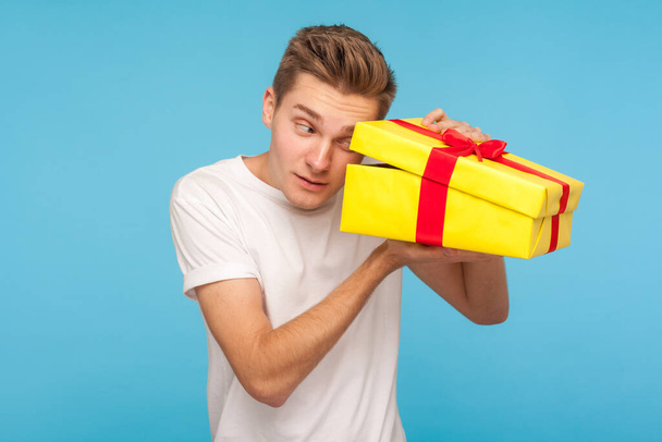 Nosy impatient man in white t-shirt looking inside gift box with curious happy expression, opening present before celebration, peeking with interest. indoor studio shot isolated on blue background - Photo, Image